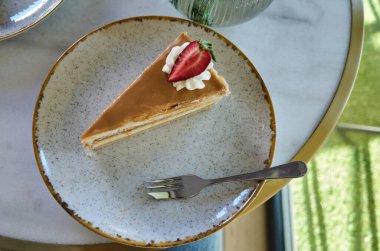 Kırsal seramik tabakta servis edilen karamel katmanlı pasta diliminin yakın çekim görüntüsü. Tatlı krem şanti ile süslenir ve üzerine küçük bir tatlı çatalı eşliğinde taze bir çilek konur. Yumuşak doğal ışık hoş ve zarif bir kafe yaratıyor.