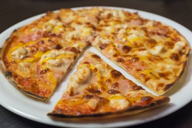 Peynirli ve jambonlu bir dilim pizza. Dilim ikiye bölünmüş. Pizza beyaz tabakta.