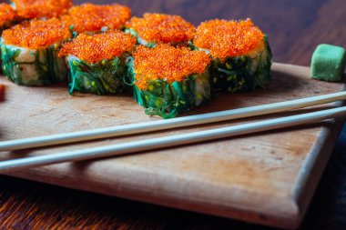 Bir tabak portakal soslu suşi ve yeşil yosun. Suşi ahşap bir kesme tahtasının üzerinde sıraya dizildi. Suşi çubuklarla servis edilir.