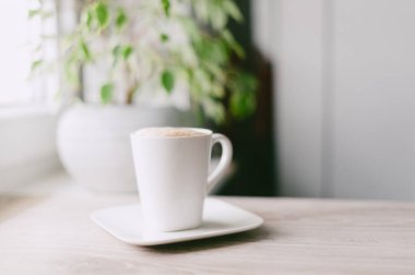 Beyaz fincan tabağı olan beyaz bir kahve fincanı ahşap bir masada oturur. Bardak kahve ile dolu ve arka planda bir bitki var.