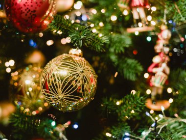 Bir Noel ağacına asılı altın bir süs. Süs diğer süslemelerle çevrili. Ağaç ışıklarla aydınlanıyor.