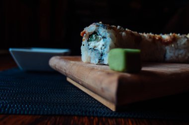 Üzerinde bir parça avokado olan suşi rulosu ahşap bir kesme tahtasının üzerinde duruyor. Yuvarlak ikiye bölünmüş ve siyah bir beze sarılmış.
