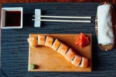 Ahşap bir tahtanın üzerinde soslu ve çubuklu bir suşi rulosu. Suşi rulosu uzun ve üzerinde bir sürü sos var.