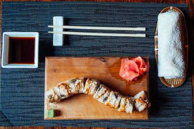 Suşi rulosu ahşap bir tahtanın üzerinde, yanında yemek çubukları ve sos var. Suşi rulosu susam tohumuyla kaplıdır ve üzerinde bir parça zencefil vardır. Sahne siyah beyaz bir masa örtüsü üzerine kurulmuş.