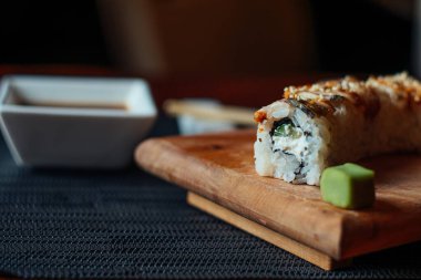 Bir suşi rulosu üzerinde küçük yeşil bir yaprak olan ahşap bir kesme tahtasının üzerinde oturur. Suşinin yanında beyaz bir kase sos var.