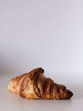 Beyaz bir arka planda bir kruvasan oturuyor. Kruvasan kahverengidir ve hafif gevrek bir dokusu vardır.