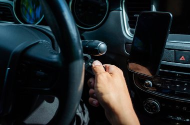 Bir erkeğin modern bir arabayı ateşleme anahtarı kullanarak çalıştırdığı yakın plan bir görüntü. Gösterge paneli, akıllı telefon montajı ve direksiyon görünür durumda. Araç işletimini, teknolojisini ve ulaşım kavramlarını gösteren yüksek çözünürlüklü fotoğraf.