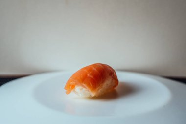 Bir parça suşi beyaz bir tabağın üzerinde duruyor. Suşi ikiye bölündü ve görüntünün ana odağı o. Tabak temiz ve dağınık, bu da suşinin ana denek olarak göze çarpmasına olanak sağlıyor.