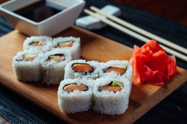 Bir tabak soslu suşi ve yemek çubuğu. Suşi kare şeklinde düzenlenir ve avokado, somon ve salatalık gibi çeşitli malzemeler içerir.