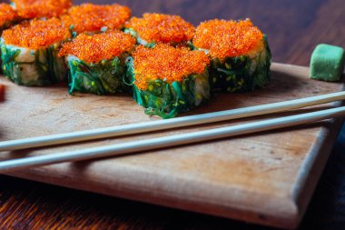 Bir tabak portakal soslu suşi ve yeşil yosun. Suşi ahşap bir kesme tahtasının üzerinde sıraya dizildi. Suşi çubuklarla servis edilir.