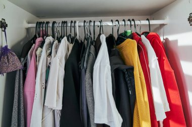 Üstünden mor bir çanta sarkan kıyafetlerle dolu bir dolap. Giysiler farklı renkte ve tarzlarda. Çanta da dolabın üstünde asılı.