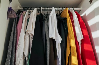 Dolapta kırmızı bir kazak ve beyaz bir gömlek var. Giysiler askıda asılı duruyor ve askıda da bir lavanta kesesi var. Sahne dağınık ve dağınık.