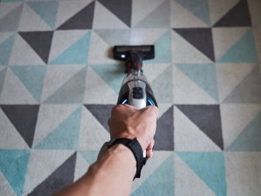 Üzerinde mavi düğme olan bir elektrikli süpürge tutuyor. Görüntünün rahat ve rahat bir havası var, çünkü kişi basit bir şekilde elektrikli süpürgeyi tutuyor ve acelesi yok.