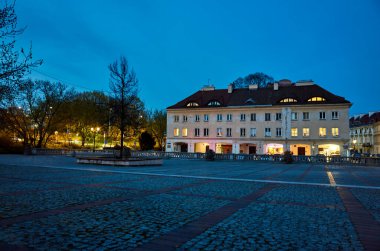 Önünde ağaç olan büyük bir bina. Bina ortasında bir çeşme olan bir avluyla çevrilidir. Avlu geceleri bomboş oluyor ve sadece birkaç ışık bölgeyi aydınlatıyor. Varşova 'daki eski kasaba. Varşova 13.04.2025 Polonya