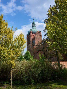 Yeşil kulesi olan yeşil ve kahverengi bir bina yemyeşil bir ormanın önünde duruyor. Bina ağaçlar ve çalılarla çevrili, huzurlu ve sakin bir atmosfer yaratıyor. Gökyüzü açık ve mavi