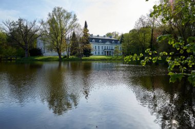 Büyük ev arka planında ağaçlar olan bir göl üzerinde yer alır. Su sakin ve gökyüzü berrak. Radziejowichi Sarayı 21.04.2025 Polonya