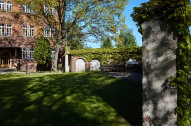 Önünde yeşil çit olan tuğla bina. Çit asmalarla kaplı. Bahçenin ortasında bir bank var.