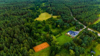 Ortasında bir ev olan yemyeşil bir orman. Evin etrafı ağaçlarla çevrili ve arka bahçesinde tenis kortu var.