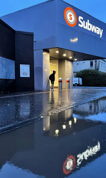 Glasgow, İskoçya 'daki Ibrox metro istasyonunun dışında ıslak bir kaldırımda tek başına duran biri. Soğuk ve nemli bir kış sabahı ve mavi ışık ışıklı turuncu metro tabelasıyla çelişiyor. Tüm sahne büyük bir su birikintisine yansıyor.