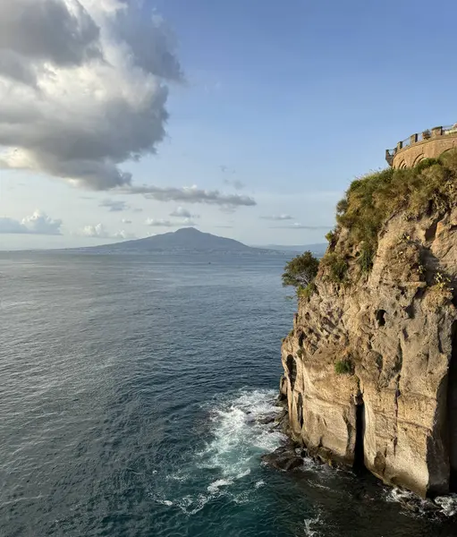 Dalgalar, Napoli Körfezi 'ndeki Sorrento kıyısındaki kayalıkların eteklerine çarpıyor. Arka planda Vezüv volkanı görünür. Mavi gökyüzünde bulutlar süzülür..