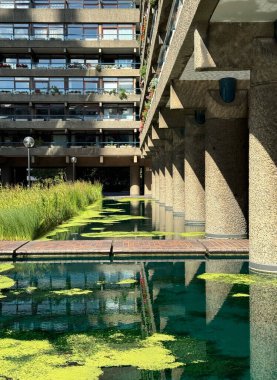 Londra 'daki Barbican' ın vahşi modernist daireleri güneşli bir günde aşağıdaki yeşil bir havuza yansıyor. Işık beton sütunlara gölge düşürür ve havuz sazlıklarla kaplıdır..