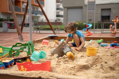 Mavi tişörtlü anaokulu çocuğu güneş gözlüğü takıyor İnşaat Oyuncaklarıyla Sandbox 'ta oynuyor.
