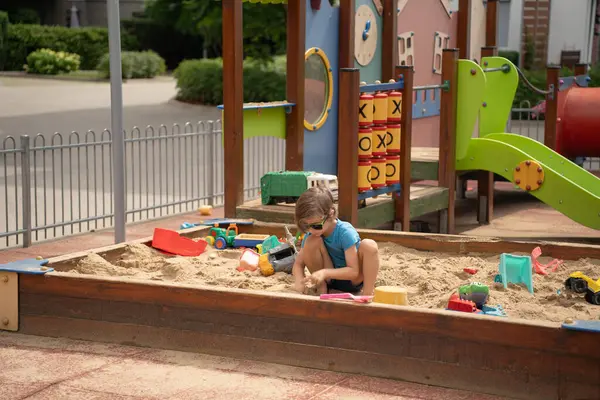 Bir çocuk şehir parkındaki bir kum havuzunda bağımsız olarak oynar. Parlak gün, kum ve plastik oyuncaklarla eğitici ve yaratıcı oyun.