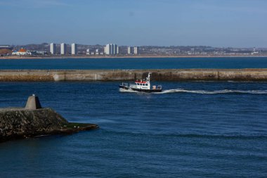 Sakin mavi sularda siyah beyaz bir teknenin dolaştığı canlı bir sahil manzarası. Arka plandaki geniş şehir manzarası botu gölgede bırakıyor. Gökdelenler de buna dahil..