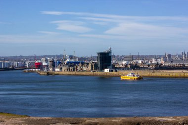 Sakin mavi sularda süzülen sarı ve mavi bir teknenin olduğu canlı bir sahil manzarası. Sanayi binaları da dahil olmak üzere, arka plandaki geniş şehir manzarası tekneyi cüce gösteriyor.