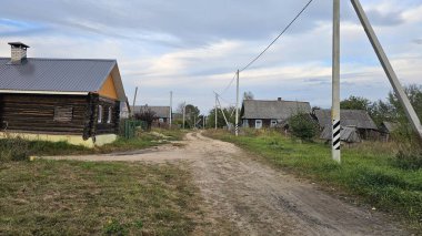 Ahşap evler Belarus köyünde bir cadde boyunca uzanan bir elektrik hattıyla çevrili. Gökyüzü bulutlu. Çimler yeşil. Rocky yolu. Bazı evler asbest kiremitlerinden yapılmıştır.
