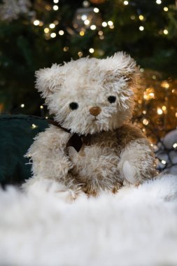 Kahverengi renkli oyuncak ayı, Noel ağacının altında beyaz bir halının üzerinde oturuyor. Ön planda beyaz halı ve arka planda Noel ışıkları.. 