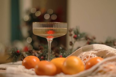 Festive Fizz. Köpüklü bir içecek dolu bir coupe kaburga bardağıyla şenlik masası. Ağ torbada mandalinalar ve arka planda çam kozalakları ve kırmızı böğürtlenlerle süslenmiş çam çelengi.