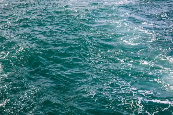 Derin deniz mavisi sularını kesen bir teknenin bıraktığı köpüklü izler. Yüzeye yayılmış köpüklü köpükler teknenin geçtiği yeri gösteriyor..