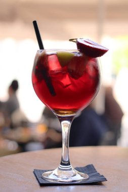 Sangria. Cam uzun bir gövdeye sahip ve tahta bir masanın üzerindeki koyu bir peçetenin üzerinde oturuyor. Parlak kırmızı bir çilek bir dekorasyon olarak camın kenarına konur. Ayrıca siyah bir pipet de var.