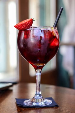 Cam uzun bir sapa sahiptir ve ahşap bir masanın üzerindeki koyu bir peçetenin üzerinde oturmaktadır. Parlak kırmızı bir çilek bir dekorasyon olarak camın kenarına konur. Ayrıca Sangria 'da siyah bir pipet var.