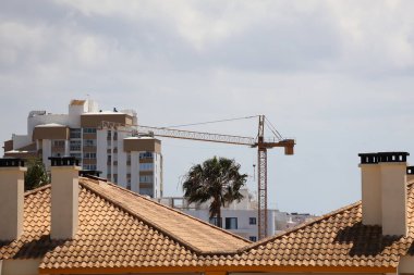 Portekiz 'in Vilamoura kentinin ikonik Akdeniz tarzı mimarisinin görüntüsü. Bu yüksek ufuk çizgisi görünümünde geleneksel kırmızı kil çatı fayansları ve konut binaları, palmiye ağaçları ve binalar arasındaki vinç üzerinde belirgin beyaz bacalar sergilenmektedir.