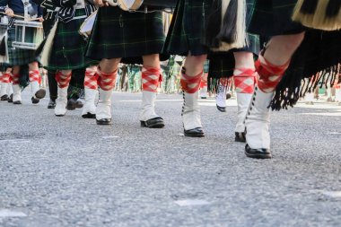 Düşük açılı bir yakın plan, geleneksel İskoç etekleri, beyaz çoraplar ve tartan çorap giyip Silahlı Kuvvetler Günü Geçit Töreni sırasında birlikte yürüyen katılımcıların bacaklarını yakalıyor. Görüntü, geçit töreninin belirgin senkronize hareketini vurguluyor.