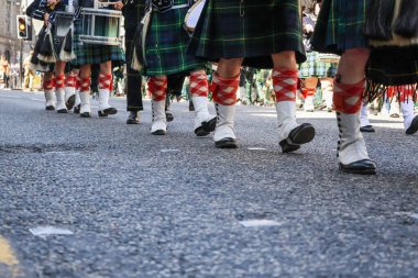 Düşük açılı bir yakın plan, geleneksel İskoç etekleri, beyaz çoraplar ve tartan çorap giyip Silahlı Kuvvetler Günü Geçit Töreni sırasında birlikte yürüyen katılımcıların bacaklarını yakalıyor. Görüntü, geçit töreninin belirgin senkronize hareketini vurguluyor.