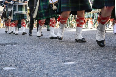 Düşük açılı bir yakın plan, geleneksel İskoç etekleri, beyaz çoraplar ve tartan çorap giyip Silahlı Kuvvetler Günü Geçit Töreni sırasında birlikte yürüyen katılımcıların bacaklarını yakalıyor. Görüntü, geçit töreninin belirgin senkronize hareketini vurguluyor.