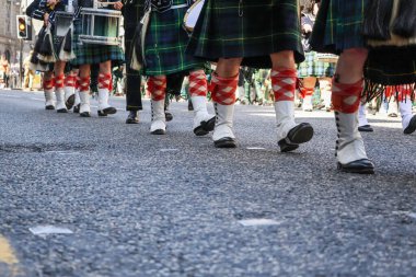 Düşük açılı bir yakın plan, geleneksel İskoç etekleri, beyaz çoraplar ve tartan çorap giyip Silahlı Kuvvetler Günü Geçit Töreni sırasında birlikte yürüyen katılımcıların bacaklarını yakalıyor. Görüntü, geçit töreninin belirgin senkronize hareketini vurguluyor.