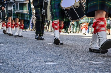 Düşük açılı bir yakın plan, geleneksel İskoç etekleri, beyaz çoraplar ve tartan çorap giyip Silahlı Kuvvetler Günü Geçit Töreni sırasında birlikte yürüyen katılımcıların bacaklarını yakalıyor. Görüntü, geçit töreninin belirgin senkronize hareketini vurguluyor.