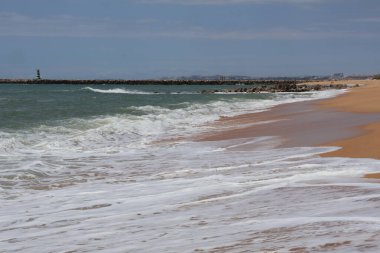 Geniş ve kumlu bir sahilde, okyanus kıyısının sakin bir görüntüsü var. Köpük desenleri ıslak kumun üzerine bırakılır ve ufukta berrak bir gökyüzünün altında sakin bir kıyı manzarası sunan uzun bir iskele uzanır.
