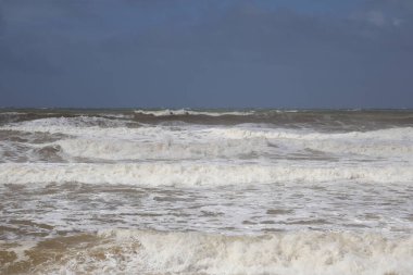 Geniş bir plajda köpüklü bir dalga çekilirken okyanus kıyısının sakin bir görüntüsü. Köpük desenleri ıslak kumun üzerine bırakılır ve ufukta berrak bir gökyüzünün altında sakin bir kıyı manzarası sunan uzun bir iskele uzanır.