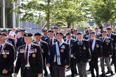 İskoçya 'nın Aberdeen kentinde düzenlenen Silahlı Kuvvetler Günü Geçit Töreni sırasında üniformalı gaziler kol geziyor. Arka plan, halkın kutlamalarının bir parçası olan disiplinli hareketlerini ve törensel başlıklarını vurguluyor.
