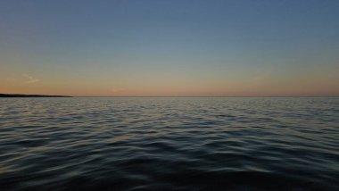 Sakin bir manzara, gün batımında sakin bir okyanusu yakalar. Suda hafif dalgalanmalar, gökyüzünün yumuşak ve sıcak tonlarını yansıtır. Ufuk çizgisi huzurlu denizi alacakaranlıktan ayırıyor, huzuru ve doğal güzelliği çağrıştırıyor.