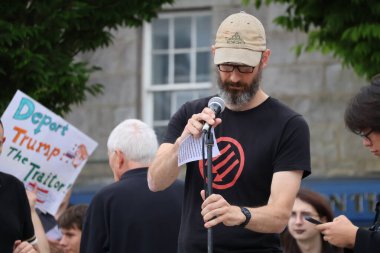 Beyzbol şapkalı ve gözlüklü sakallı bir adam Aberdeen 'deki bir protestoda mikrofona tutkuyla konuşuyor. Arkaplanda bulanık figürler ve şehir mimarisi ile mesajının ciddiyetini aktarıyor.