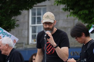 Beyzbol şapkalı ve gözlüklü sakallı bir adam Aberdeen 'deki bir protestoda mikrofona tutkuyla konuşuyor. Arkaplanda bulanık figürler ve şehir mimarisi ile mesajının ciddiyetini aktarıyor.