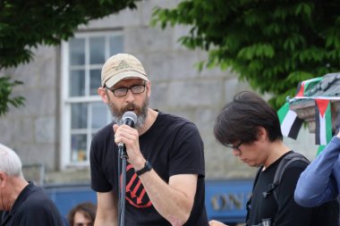 Beyzbol şapkalı ve gözlüklü sakallı bir adam Aberdeen 'deki bir protestoda mikrofona tutkuyla konuşuyor. Arkaplanda bulanık figürler ve şehir mimarisi ile mesajının ciddiyetini aktarıyor.