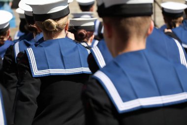 Arkadan yakından bakınca, koyu mavi üniformalı, düz şapkalı, lacivert üniformalı birçok kişi görülüyor. Görünür yüzleri veya özel işaretleri olmayan başları ve omuzlarına verilen ilgi, birlik ve disiplini vurguluyor..