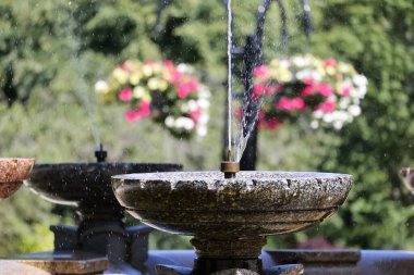 İki taş çeşme, biri benekli gri granitten diğeri ise sıcak kırmızımsı kahverengi taştan yapılmış, huzurlu bir parkta su püskürtüyor. Uzun su püskürtüsü ve yeşil ağaçlar ve yaprakların bulanık arkaplanı dingin ve ferahlatıcı bir manzara yaratır.